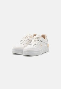 Zapatillas blancas con un upper de cuero liso, cuello acolchado y una suela blanca gruesa. Presenta un acento dorado en el talón y cordones en tono.