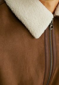 Giacca in suede marrone con colletto in sherpa bianco e zip frontale, in primo piano sulla parte superiore del petto e dell'area del collo.