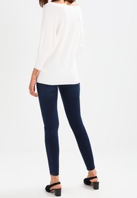 Weißer, strukturierter Pullover mit weitem Ausschnitt, kombiniert mit dunkelblauen Skinny-Jeans und schwarzen Sandalen mit Blockabsatz. Von hinten gestylt.