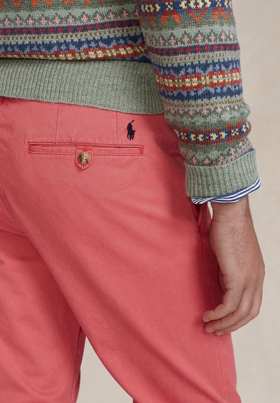 Πλεκτό πράσινο πουλόβερ με πολύχρωμα σχέδια· κοραλλί παντελόνια με πίσω τσέπη με κουμπί και μικρό κεντημένο λογότυπο. Ορατά ριγέ μανσέτα.