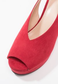 Chaussure à talon haut en daim rouge à bout ouvert avec doublure intérieure beige et coutures visibles sur fond blanc.