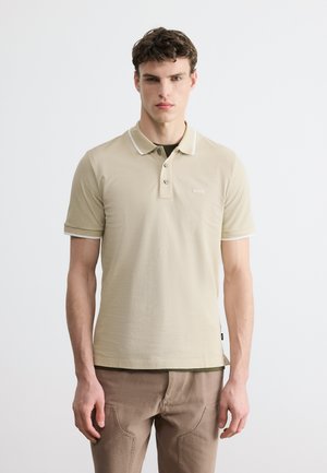 Νεαρός άνδρας φοράει μπεζ πόλο πουκάμισο με λευκή επένδυση και καφέ παντελόνι, στέκεται μπροστά σε απλό φωτεινό φόντο.