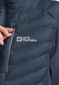 Dunkelblauer, isolierter Weste mit gestepptem Design, Reißverschluss-Tasche und grauem Logo "Jack Wolfskin" auf der Vorderseite. Glatte Textur.