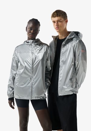Két modell fényes ezüst kapucnis dzsekit visel, az egyik zipzárosra húzva, a másik nyitva, mindkettő fekete ruházattal alatta.
