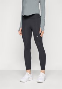 Leggings neri realizzati in tessuto elastico, con un design aderente e un piccolo logo bianco Nike sulla coscia sinistra. Indossati con una maglietta grigia e scarpe bianche.