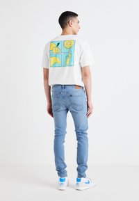Camiseta blanca de algodón con un colorido gráfico de limones en la parte posterior, combinada con jeans azul claro y zapatillas blancas con acentos azules.