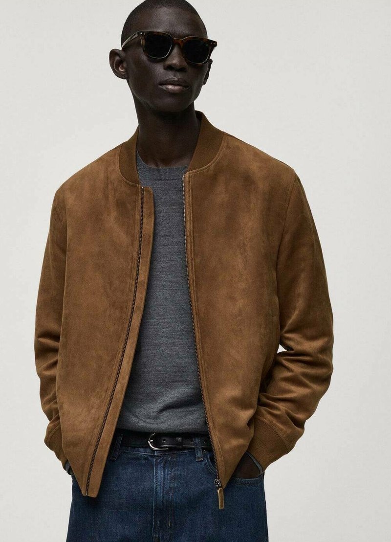 Giubbotto bomber in suede marrone, colletto a costine, chiusura con zip, abbinato a un maglione grigio e jeans in denim scuro. Finitura opaca; senza modelli visibili.