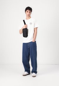 Weißes T-Shirt mit Grafik-Logo, lässige blaue Jeanshose und kleine schwarze Umhängetasche mit Reißverschluss. Leichte Sneakers runden das Outfit ab.