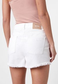 Shorts di denim bianco con orlo sfrangiato, due tasche posteriori e una etichetta in pelle marrone con la scritta "DENIM". Il tessuto sembra robusto e spesso.