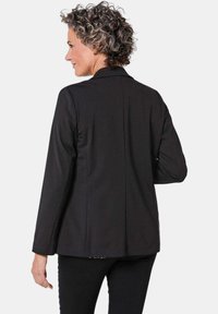 Zwarte blazer met een getailleerde pasvorm, gelapte revers, lange mouwen en een gladde stofafwerking. Aan de achterkant bevindt zich een split voor extra bewegingsvrijheid.