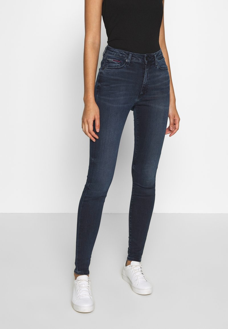 Jeans ajustados en denim de lavado oscuro con un sutil desgastado, cintura de altura media, diseño de cinco bolsillos, combinados con zapatillas blancas.