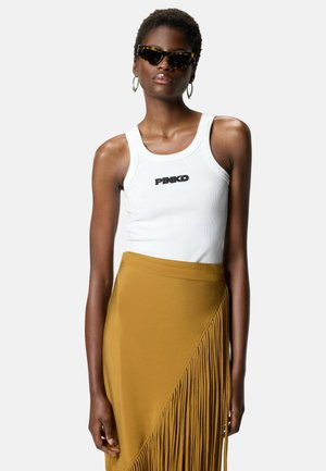 Donna con occhiali da sole tartarugati, orecchini a cerchio, top bianco senza maniche con logo "PINKO" e gonna color senape con dettagli di frange.