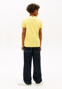 Αγόρι με σγουρά μαλλιά που φοράει κίτρινο μπλουζάκι, σκούρα άνετα τζιν με μεγάλες πίσω τσέπες και λευκά αθλητικά, στέκεται με την πλάτη γυρισμένη.