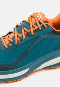Tealové a oranžové sportovní boty s texturovaným svrškem, integrovaným šněrovacím systémem, polstrovaným límcem a kontrastními bílými a oranžovými detaily na podrážce.