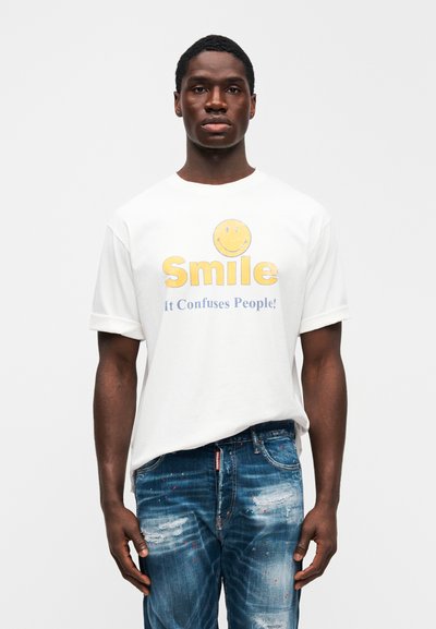 Λευκό βαμβακερό μπλουζάκι με κίτρινο γραφικό με χαμογελαστό πρόσωπο και κείμενο που γράφει «Smile It Confuses People!» Συνδυάζεται με τζιν παντελόνι σε μπλε χρώμα με φθορές.