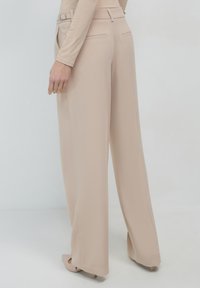 Pantalons larges beiges en tissu lisse, dotés de deux poches arrière et d'une taille plate avec des accents en métal.