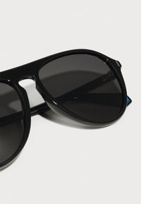 Ochelari de soare negri cu un design elegant și rotunjit, având lentile întunecate polarizate. Prezintă un finisaj lucios și o branduire minimă pe rame.
