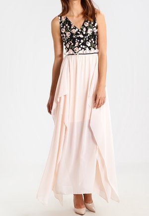 Mouwloze jurk met een zwart geborduurde top met bloemen en een vloeiende asymmetrische rok in lichtroze, gedragen met naaldhakken in nudekleur.