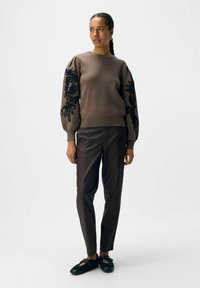 Brązowy sweter z czarnym haftem kwiatowym na rękawach, zestawiony z ciemno-brązowymi spodniami i czarnymi butami na slidery. Miękka tkanina, luźny krój.