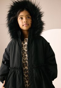 Black puffer coat with a black faux fur hood, zipper front, and gathered waist, worn over a leopard print sweater. Smooth fabric texture.