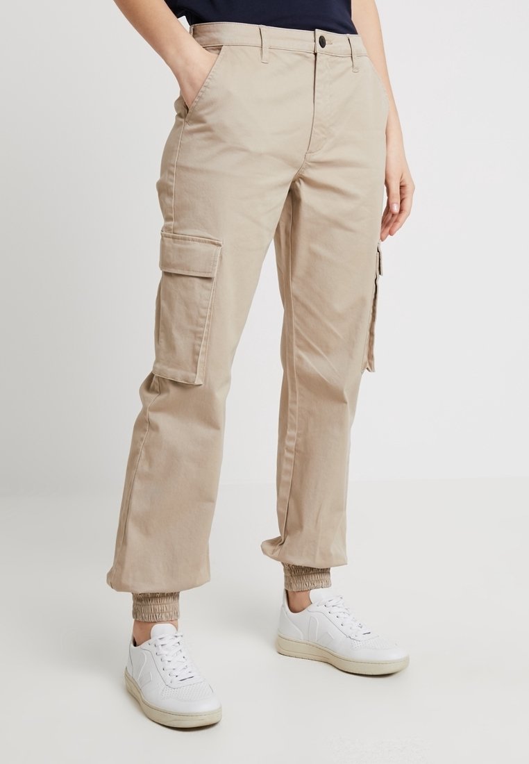 Pantalon cargo beige avec poches à rabat latérales et poignets élastiques, porté avec des baskets blanches et un haut foncé, modèle debout les mains dans les poches.