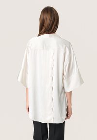 Camicia di colore crema, morbida e fluida, con maniche corte e un design a scallop lungo la cucitura posteriore.
