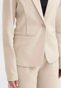 Beige op maat gemaakte blazer van gladde stof, met een enkele knoopsluiting, notch-revers, twee voorzakken en lange mouwen.