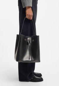 Mustast nahast ämbrikujuline kott tõmblukuga, sileda tekstuuri ja kuldse pealispinnaga. Stiilselt kombineeritud tume teksapükste ja mustade kingadega.