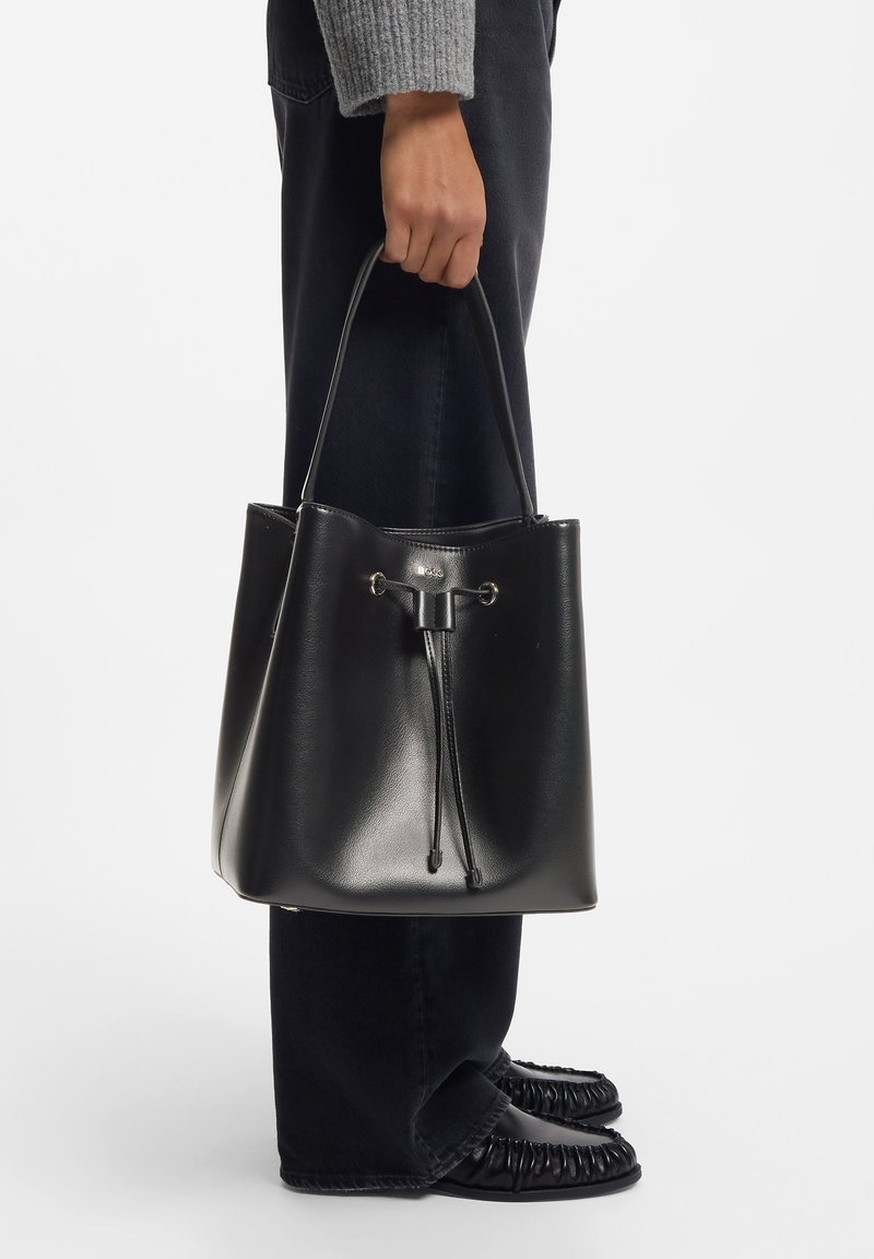 Mustast nahast ämbrikujuline kott tõmblukuga, sileda tekstuuri ja kuldse pealispinnaga. Stiilselt kombineeritud tume teksapükste ja mustade kingadega.