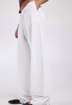 Person wearing loose white pants with scalloped hem and black slip-on shoes, standing with one hand in pocket.