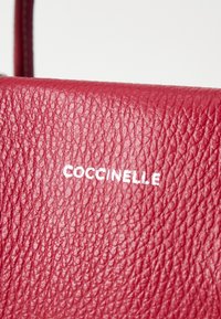 Rdeče usnje z teksturirano površino. Ima vtisnjeno blagovno znamko "Coccinelle" v beli barvi. Kompakten, pravokoten dizajn.