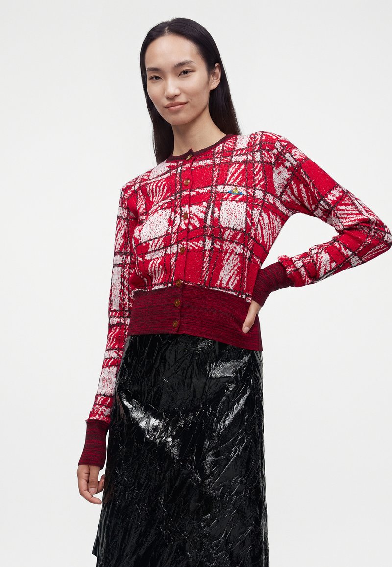 Femme portant un cardigan boutonné à motif rouge et blanc et une jupe noire texturée brillante, debout avec une main sur la hanche.