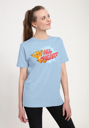 Młoda kobieta z kucykiem, ubrana w jasnoniebieską koszulkę z uśmiechniętym pomarańczowym misiem i czerwonym napisem „All Heart”, patrząca w górę, stojąca.