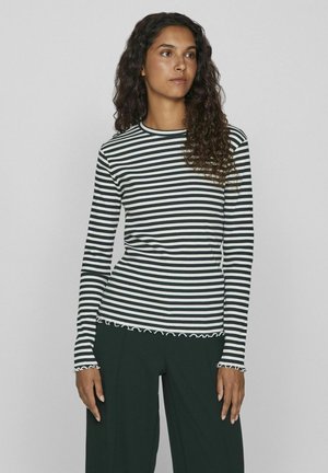Top de rayas en blanco y negro de manga larga con cuello redondo y dobladillo con volantes. Textura de canalé suave; silueta ajustada. Combinado con pantalones verde oscuro.