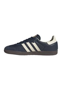 Sneaker blu navy con tre strisce beige, lacci neri, linguetta beige sul tallone e suola marrone texturizzata, vista laterale.