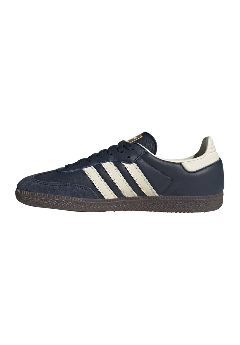 Sneaker blu navy con tre strisce beige, lacci neri, linguetta beige sul tallone e suola marrone texturizzata, vista laterale.