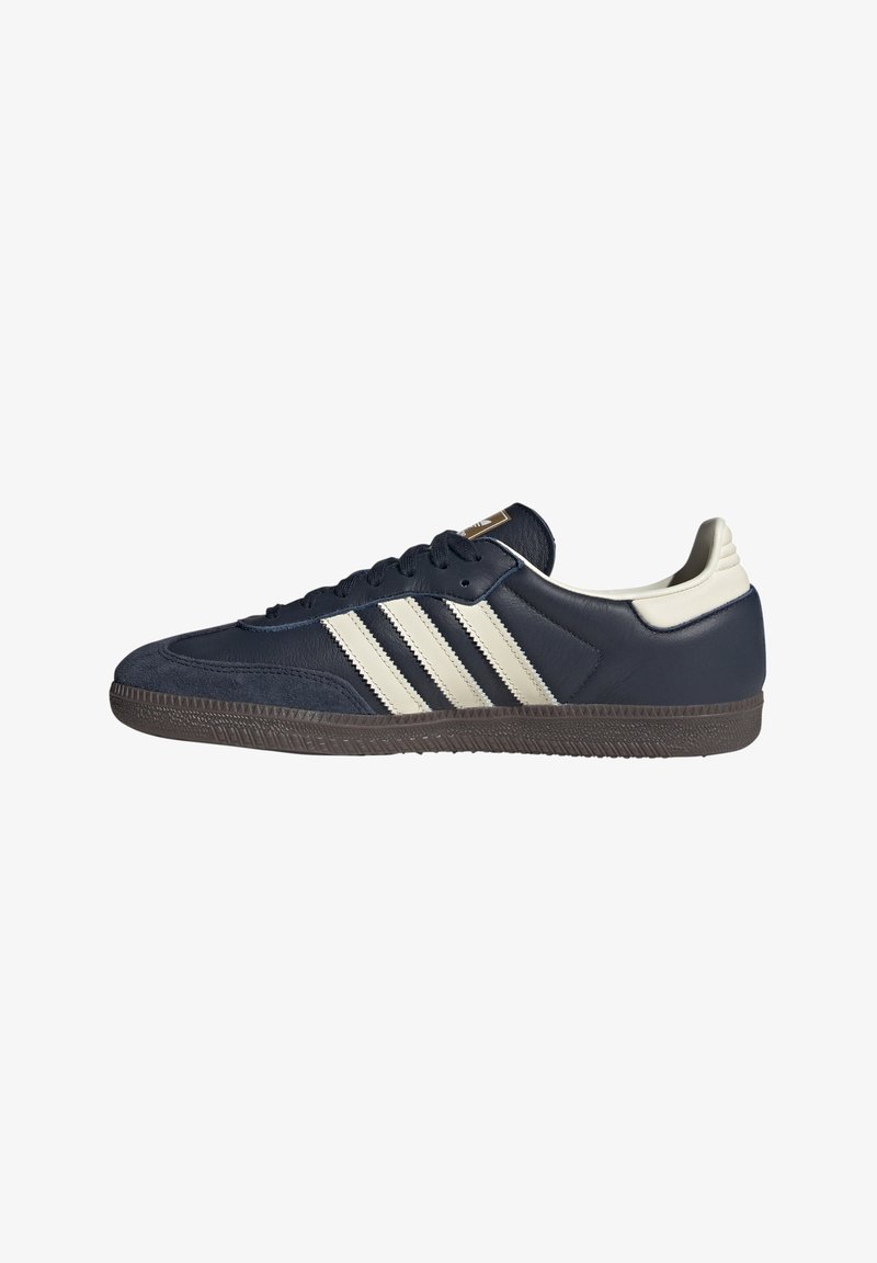 Sneaker blu navy con tre strisce beige, lacci neri, linguetta beige sul tallone e suola marrone texturizzata, vista laterale.