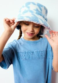Chapeau en polaire bleu clair avec un motif d'étoiles blanches, porté de travers. T-shirt bleu arborant "NEW YORK" en broderie texturée.