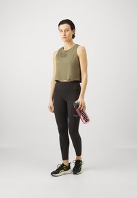 Oliivroheline lühike tank top, mustad kõrge vöökohaga trennipüksid, mustad tennisetossud roheliste detailidega, hoides roosa veepudelit musta tekstiga.