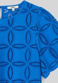 Blouse bleue en tissu texturé avec des motifs floraux découpés et un col arrondi. Manches courtes avec une coupe décontractée. Taille S.