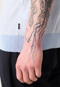 Maglione lavorato a maglia blu chiaro con orlo a coste, dotato di un'etichetta con logo. Primo piano di una mano poggiata su un tessuto scuro, che mette in evidenza un tatuaggio dettagliato.