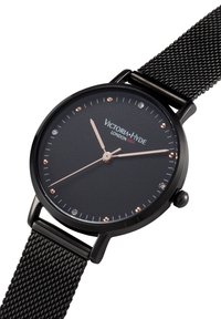 Okrągły czarny zegarek z matową, teksturowaną tarczą, różowo-złotymi wskazówkami, kryształowymi znacznikami godzin oraz czarnym paskiem siateczkowym. Nazwa marki wyraźnie widoczna.