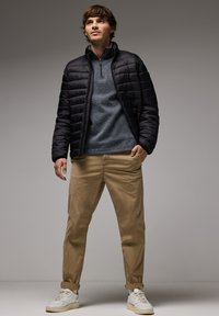 Giacca a vento nera trapuntata sopra un pullover grigio scuro a maglia, abbinata a chinos beige e sneaker bianche, in piedi su uno sfondo neutro.