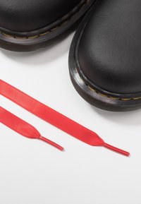 Černé kožené boty s žlutým šitím a nerozvázanými červenými plochými tkaničkami umístěné na bílé ploše.