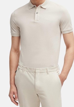 Mann trägt hellbeiges kurzärmeliges Polohemd und passende Hose mit einer Hand in der Tasche vor einfarbigem Hintergrund.