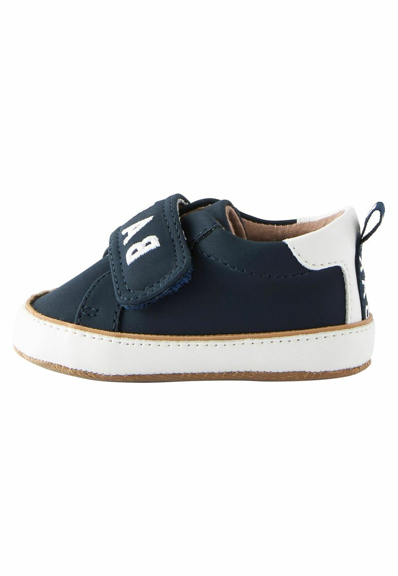 Chaussure en tissu bleu marine avec des accents blancs, dotée d'une sangle Velcro et d'un bout arrondi. Semelle en caoutchouc avec un bord beige et un petit onglet à l'arrière.