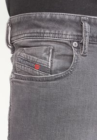 Närbild av en grå denimjeansficka med synlig bältesöggla, sömmar och en liten varumärkesetikett i svart och rött.