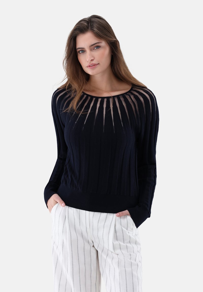 Frau mit langen Haaren, die einen schwarzen Pullover mit durchscheinendem, gestreiftem Einsatz und eine weiße Hose mit dünnen vertikalen Streifen trägt, die Hände in den Taschen.