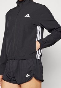 Sort atletisk jakke med høj krave, fuld lynlås og hvidt logo; parret med sorte shorts med hvide detaljer og side-striber.