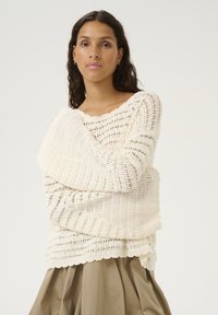 Stickad tröja i krämfärg med en öppen, texturerad design. Har en vågig halsringning och ribbad nederdel, tillsammans med en beige kjol.